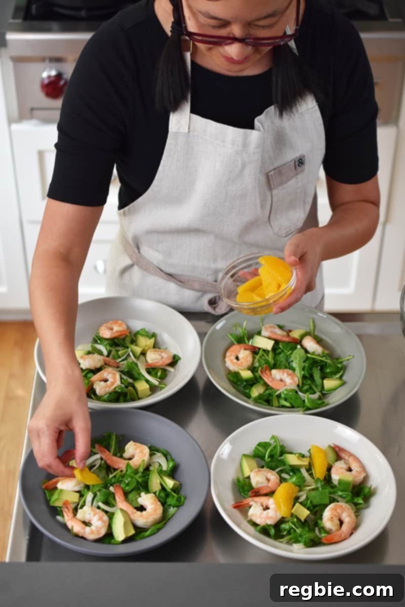 An Asian woman in a beige apron is adding orange segments to four bowls of shrimp and avocado salad