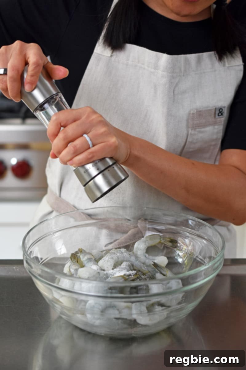 A person in a beige apron is adding freshly cracked black pepper to a bowl of peeled raw shrimp.