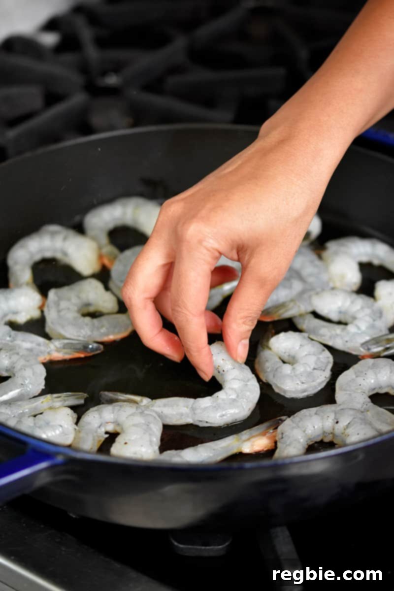 A hand is adding seasoned raw shrimp to a large cast iron skillet on the stovetop.