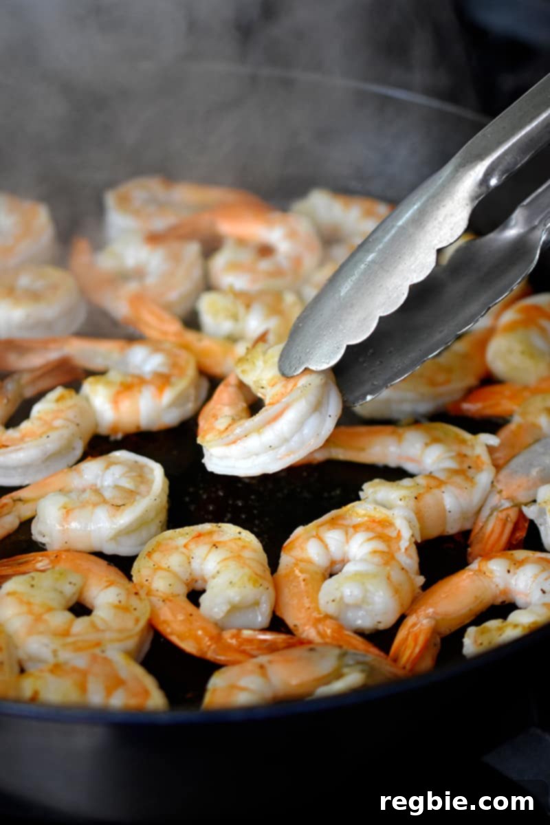 A pair of tongs is flipping shrimp in a large cast iron skillet.