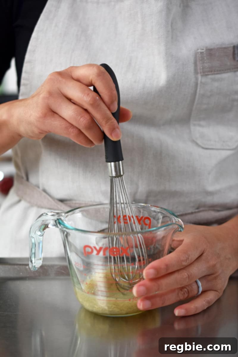 A person in a beige apron is whisking champagne vinaigrette in a liquid measuring cup.