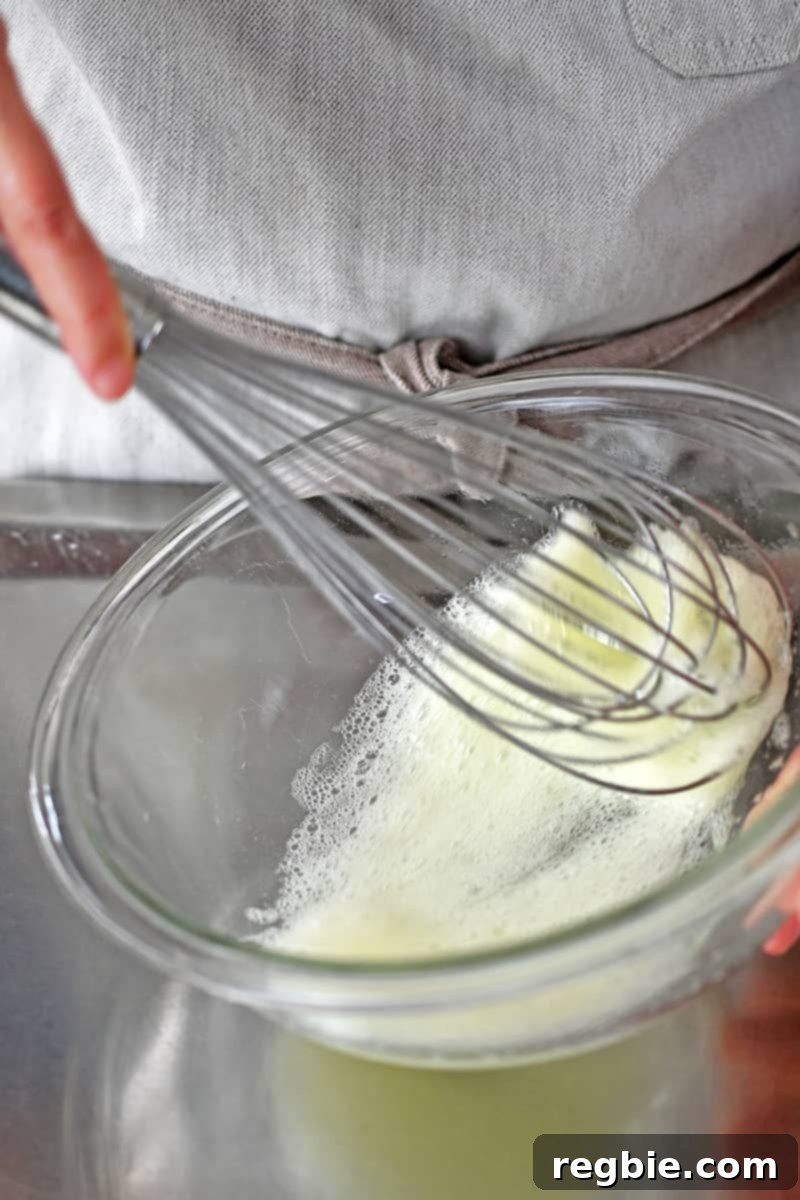 A whisk is beating egg whites until they are frothy in a glass bowl, creating the binder for the calamari coating.