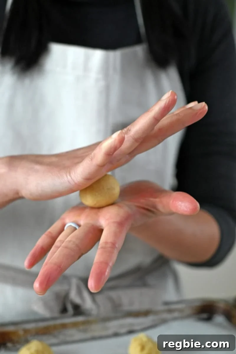 Hands are shown gently rolling a portion of cookie dough into a smooth, 1-inch ball.