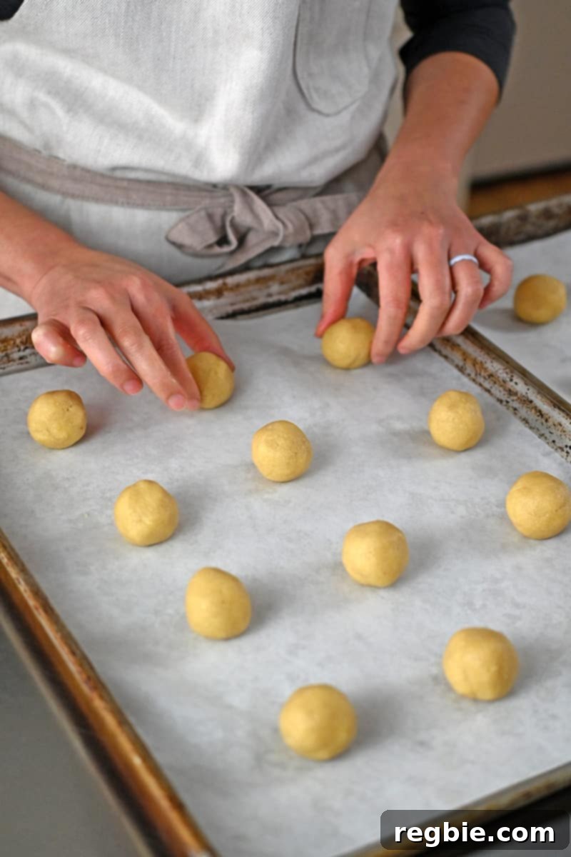 Two hands are meticulously arranging small balls of cookie dough onto a parchment-lined rimmed baking sheet.