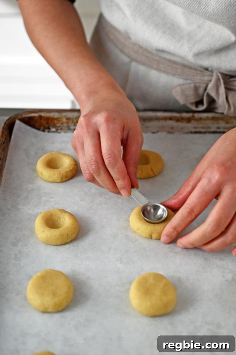 Someone is expertly using a half-teaspoon measuring spoon to create a neat indentation in a flattened cookie dough ball.