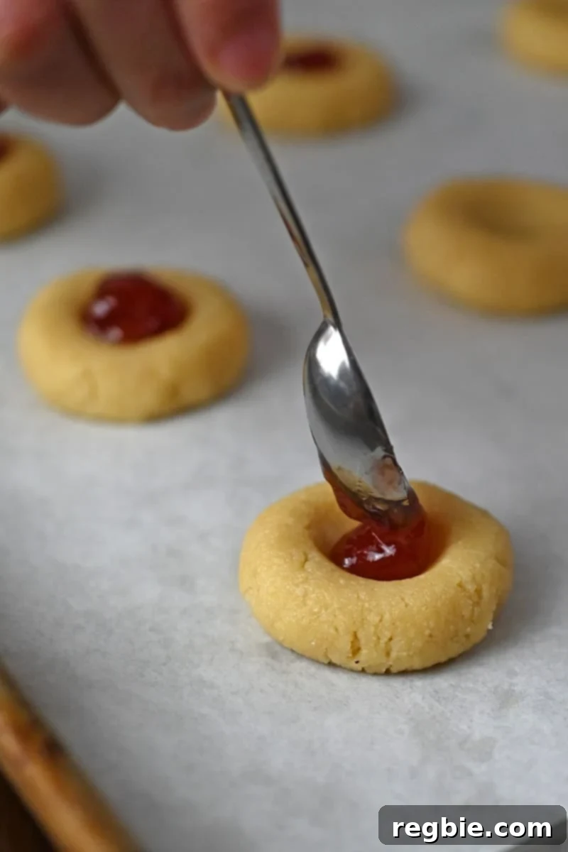 A small spoon is delicately adding a dollop of vibrant jam to the hollowed center of a thumbprint cookie dough.