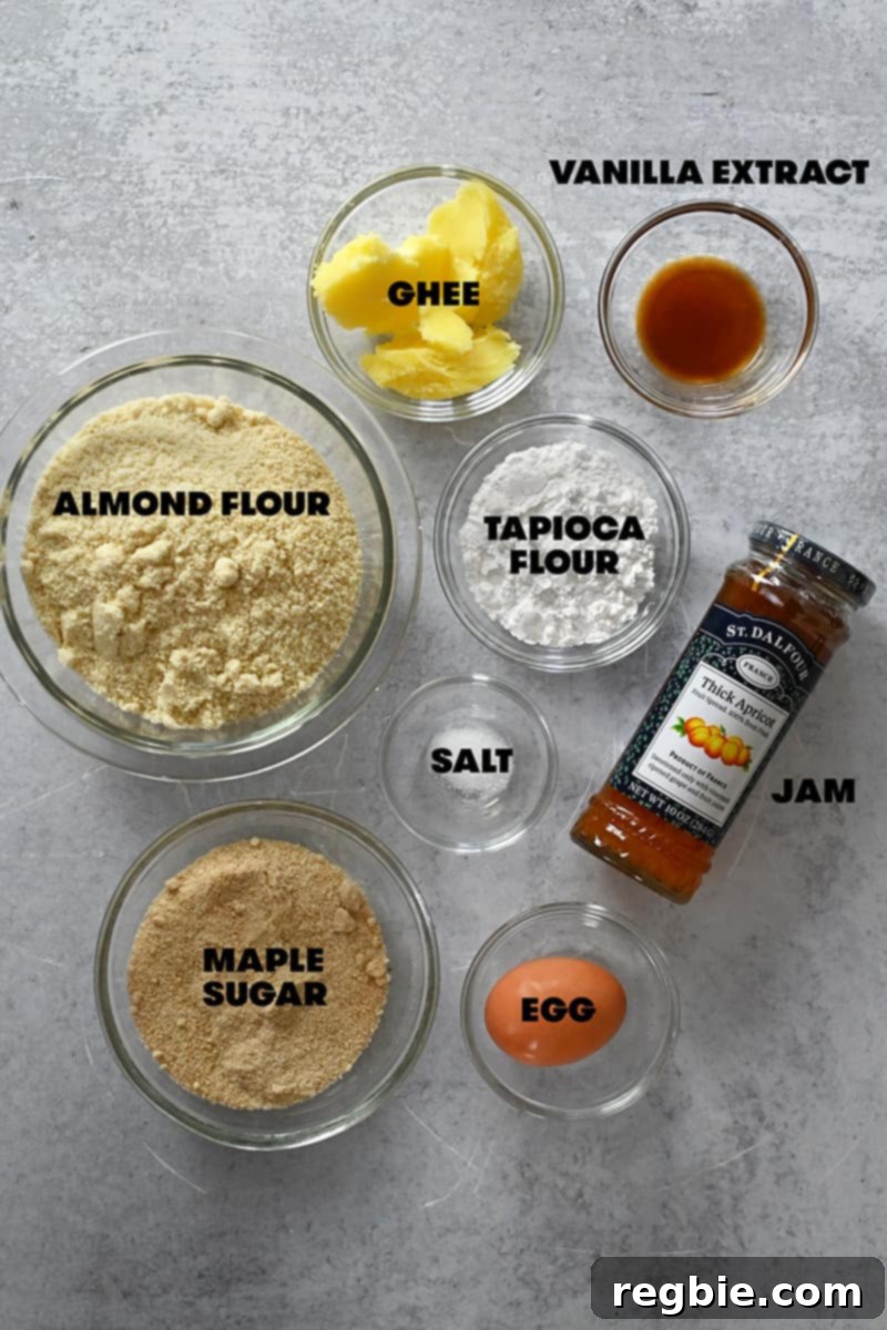 An overhead shot of the raw, whole ingredients laid out for making paleo and gluten-free thumbprint cookies: various flours, sweeteners, fats, eggs, and jars of jam.