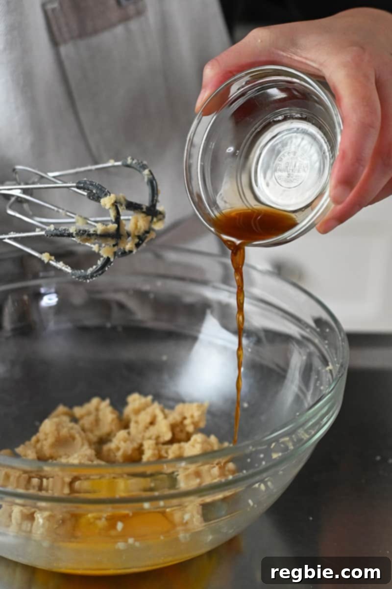 Vanilla extract is poured into a bowl containing creamed sugar, ghee, and a raw egg, ready to be mixed.