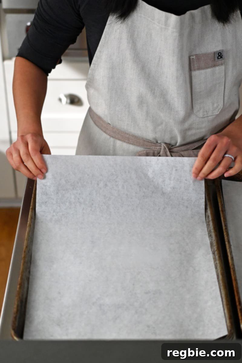 A person in an apron carefully places a piece of parchment paper onto a rimmed baking sheet, preparing it for baking.