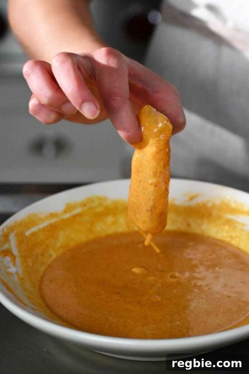 A hand is carefully dipping a fish fillet into a shallow bowl containing the seasoned egg and cassava flour batter, ensuring an even coating.