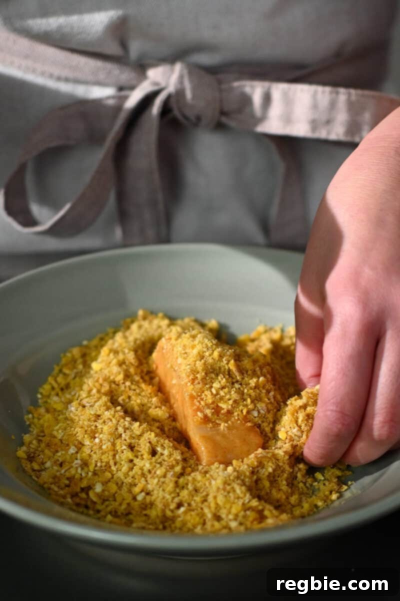 A fish fillet is nestled in a shallow dish filled with golden plantain chip crumbs, being gently pressed to ensure full coverage.