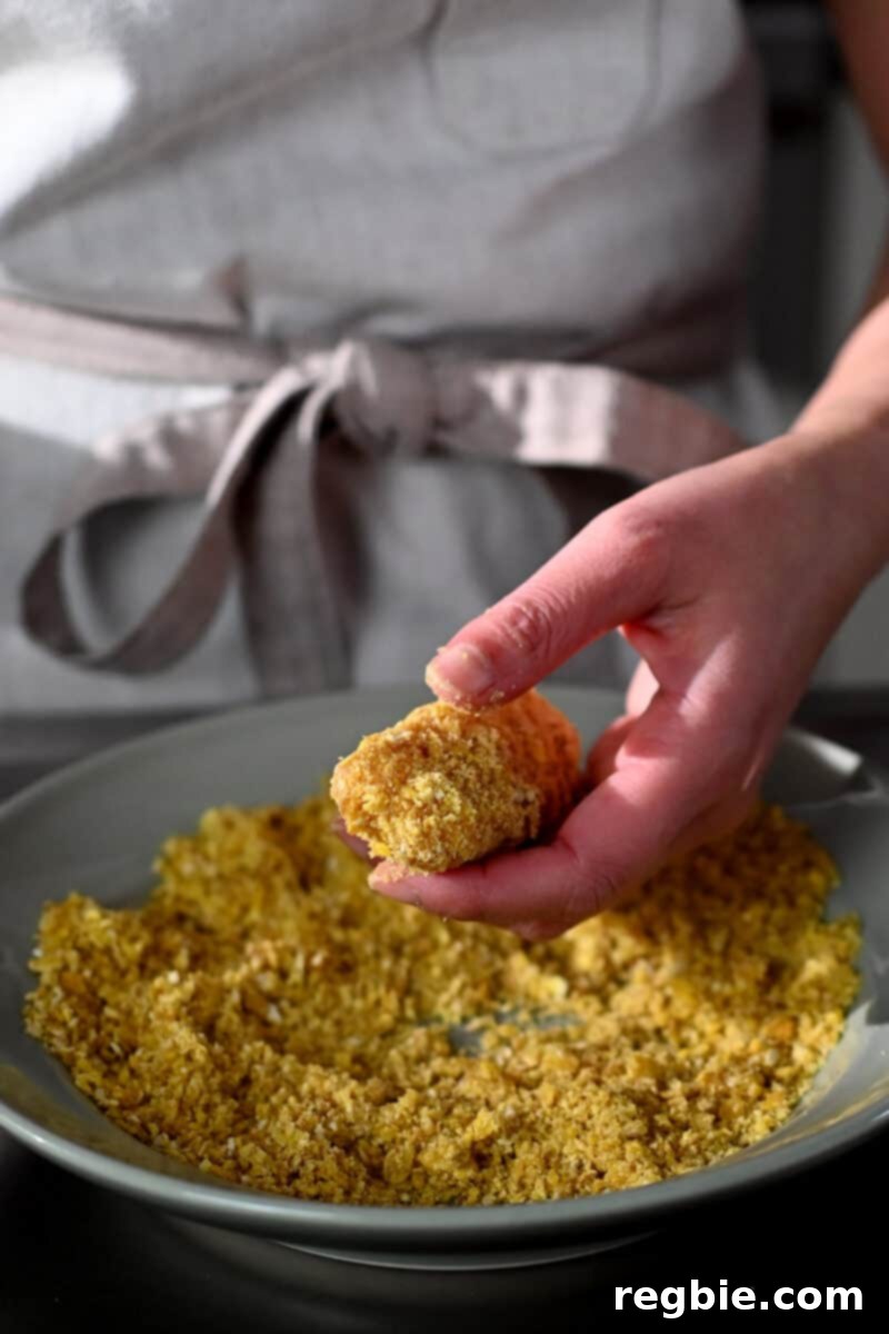A hand is meticulously coating a fish stick with plantain chip crumbs, ensuring full and even coverage before cooking.