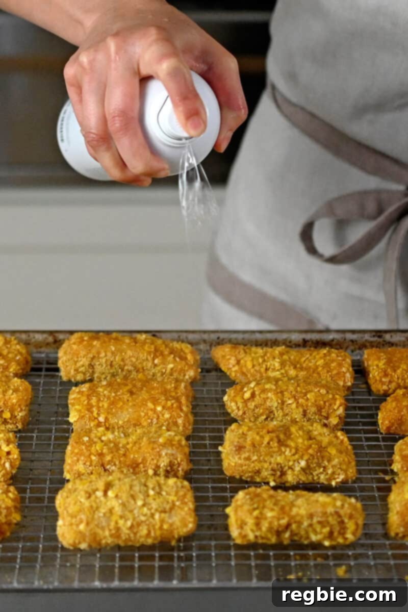 A hand is spraying a wire rack covered with uncooked fish sticks with avocado oil spray, preparing them for the air fryer.