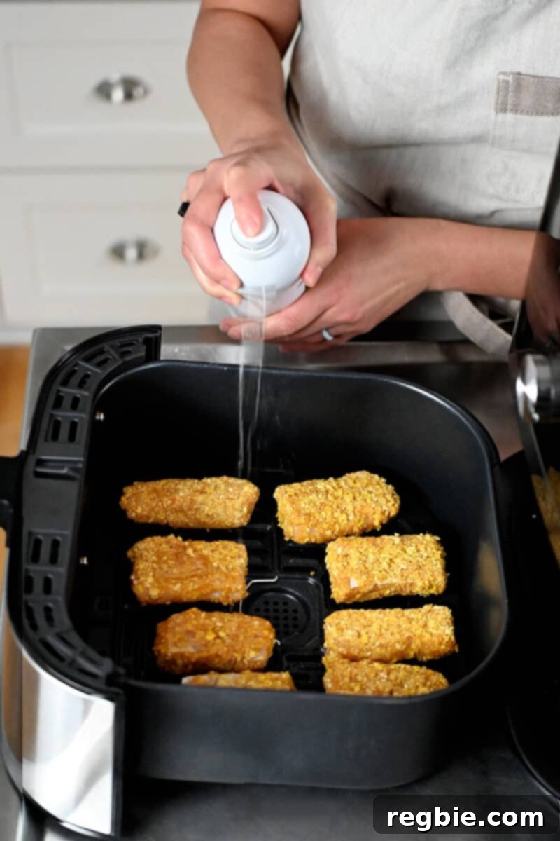 A hand is spraying avocado oil onto fish sticks neatly arranged in a single layer within an air fryer basket, ensuring even cooking.