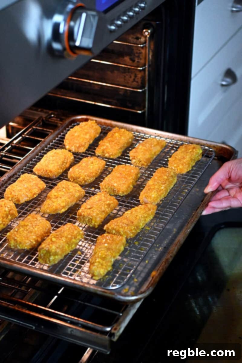 A hand is carefully placing a tray of paleo fish sticks, arranged on a wire rack within a baking sheet, into a preheated oven.
