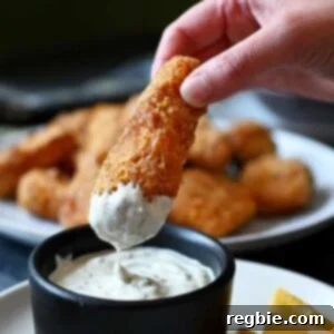 A hand is dipping a crispy homemade fish stick in creamy tartar sauce, showcasing its golden-brown crust.