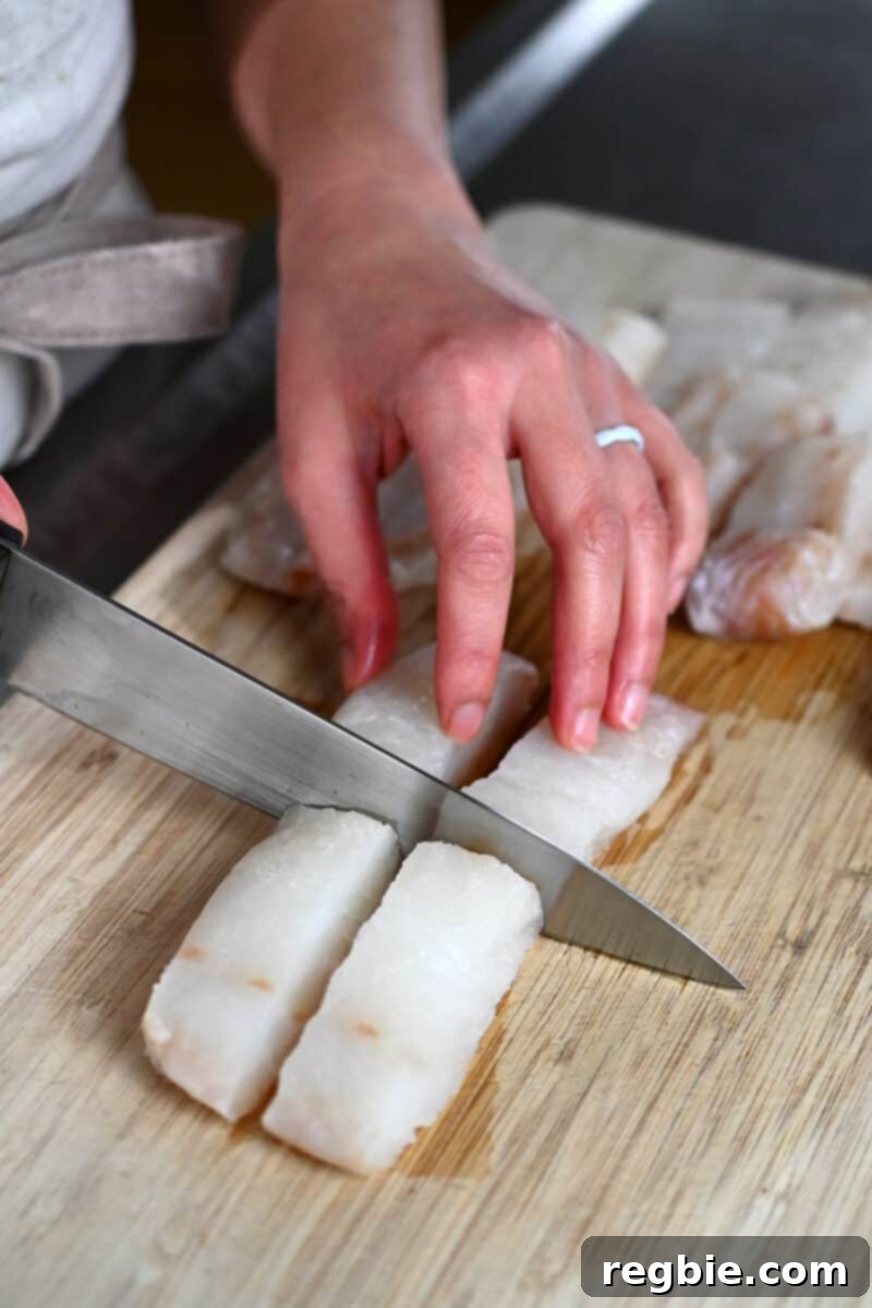 A boneless and skinless cod fillet is being precisely cut into four uniform 3x1-inch pieces on a cutting board, preparing for homemade fish sticks.