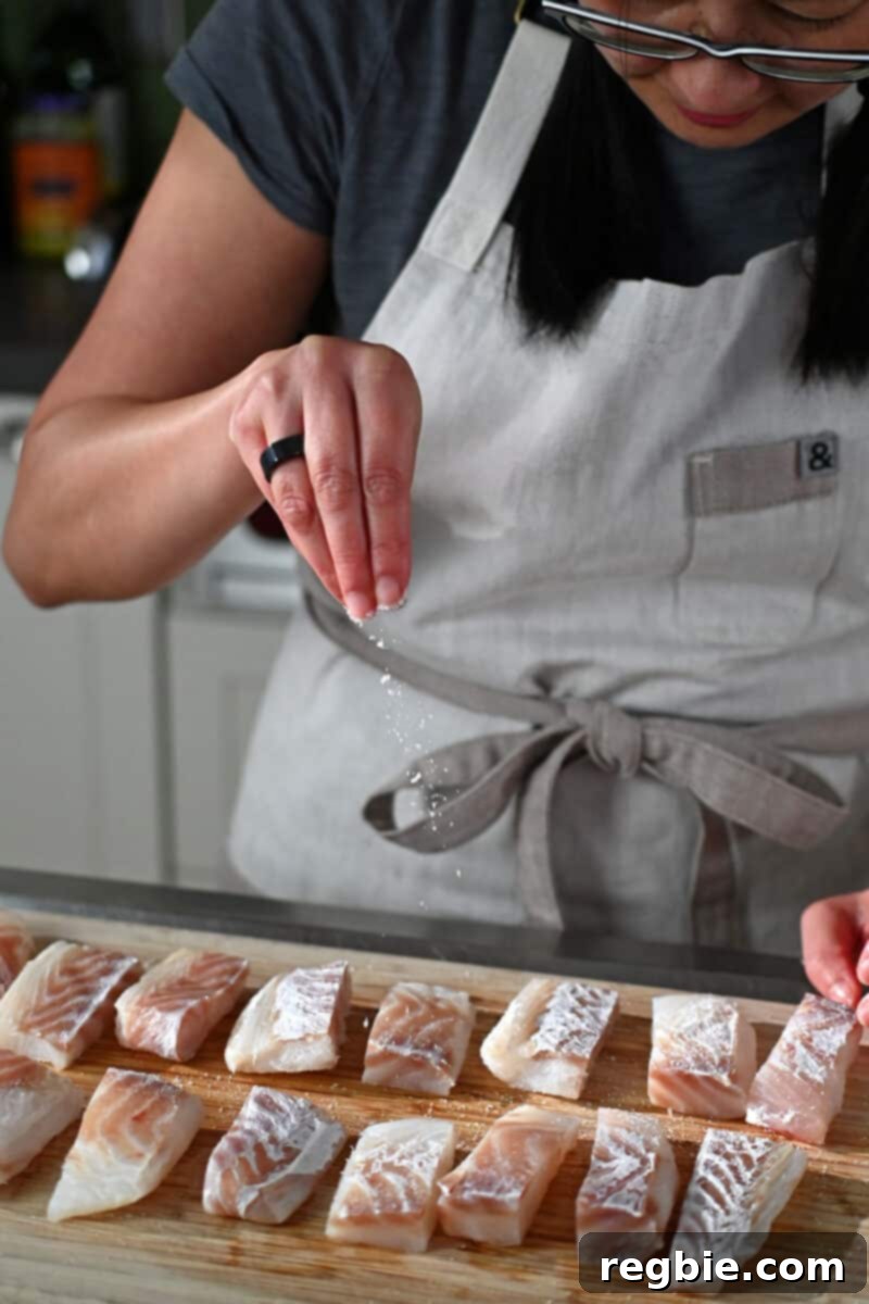 A hand is sprinkling Diamond Crystal kosher salt evenly over cod fish sticks arranged on a wooden cutting board, preparing them for battering.
