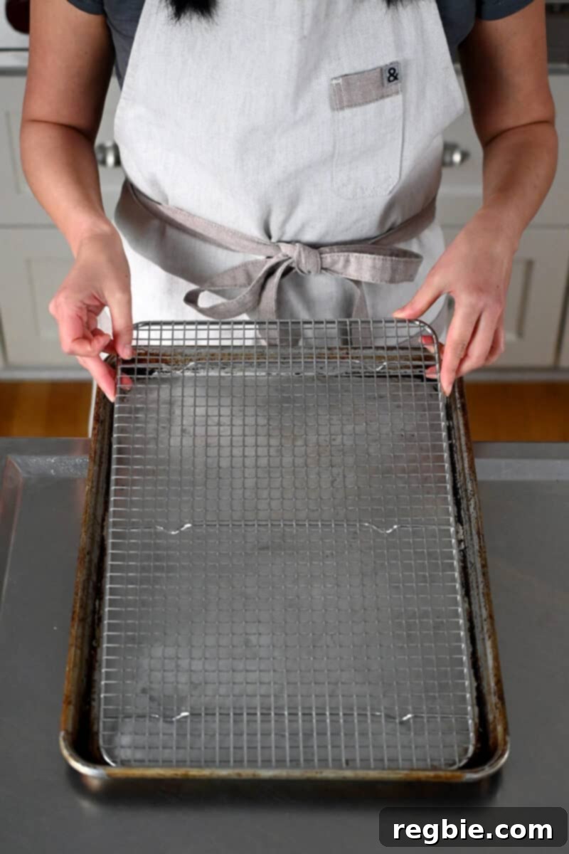 A person in an off-white apron is carefully placing a silver wire rack inside a black rimmed baking sheet, setting up a dredging station.