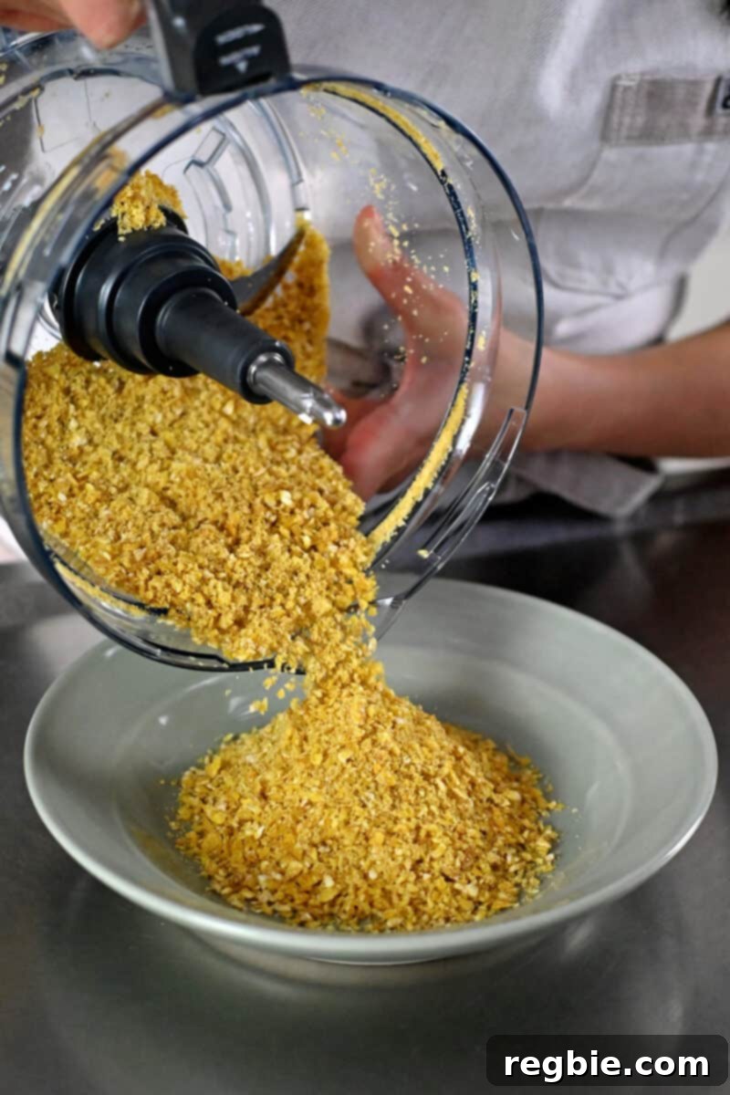 A hand is pouring half of the finely processed plantain chip crumbs into a shallow green bowl, preparing for the coating process.
