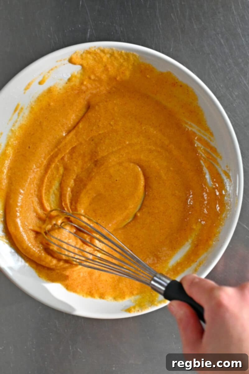 An overhead shot of a whisk combining eggs, cassava flour, and a blend of spices in a shallow dish, forming a uniform sticky paste for the fish stick batter.