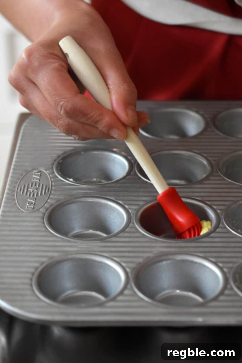 Paleo Raspberry Financiers 5 A hand meticulously uses a silicone brush to apply melted ghee, greasing the individual cups of a mini muffin tin, ensuring a non-stick surface for baking.