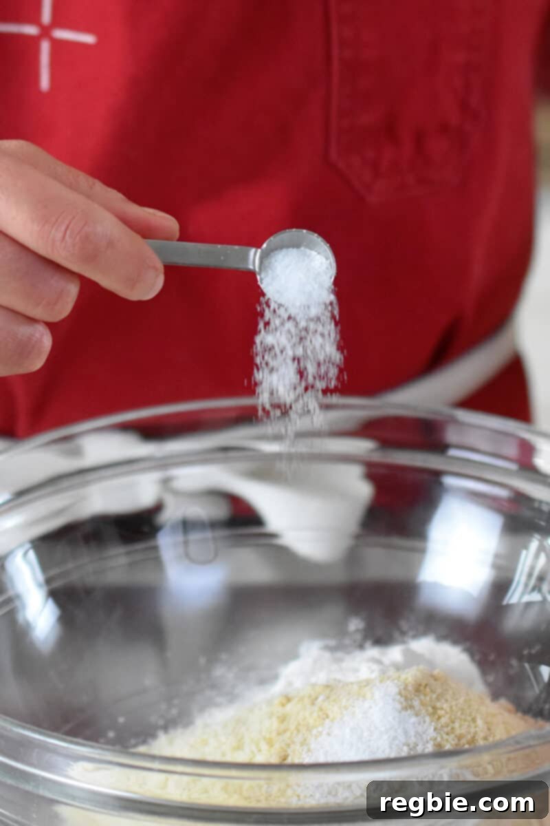Paleo Raspberry Financiers 6 A close-up shot of a hand adding a spoonful of sparkling Diamond Crystal kosher salt into a clear glass bowl, already containing the almond flour, maple sugar, and cassava flour for the paleo financiers.