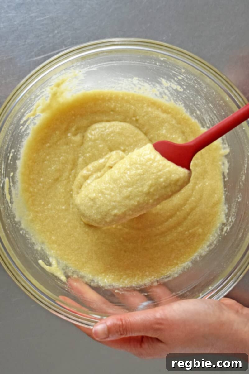 Paleo Raspberry Financiers 8 A bright red spatula smoothly folds and mixes the paleo financier batter in a glass mixing bowl, showcasing its uniform and rich consistency.