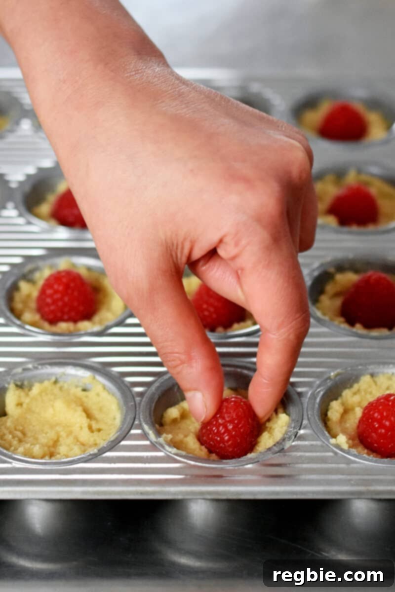 Paleo Raspberry Financiers 10 A delicate hand carefully places a fresh, plump raspberry into the center of the financier batter in a mini muffin pan, preparing it for baking.