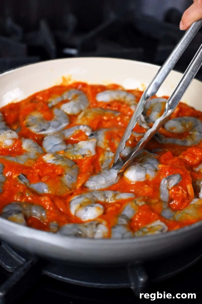 Adding large peeled shrimp into a skillet with spicy marinara sauce, ensuring they are evenly distributed.