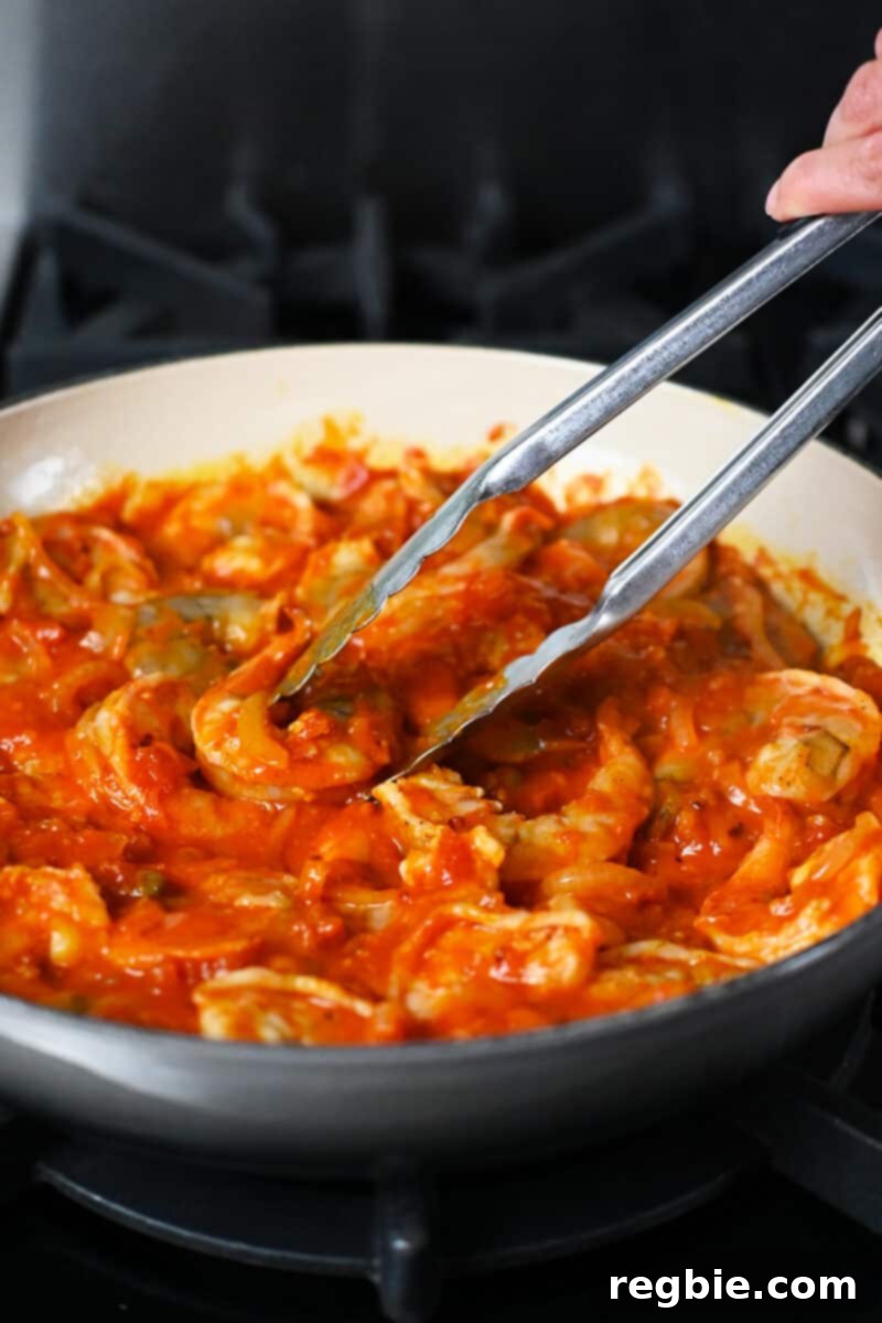 A pair of tongs are picking up fully cooked shrimp in an enamel cast iron skillet with spicy marinara sauce, showing their perfectly pink color.