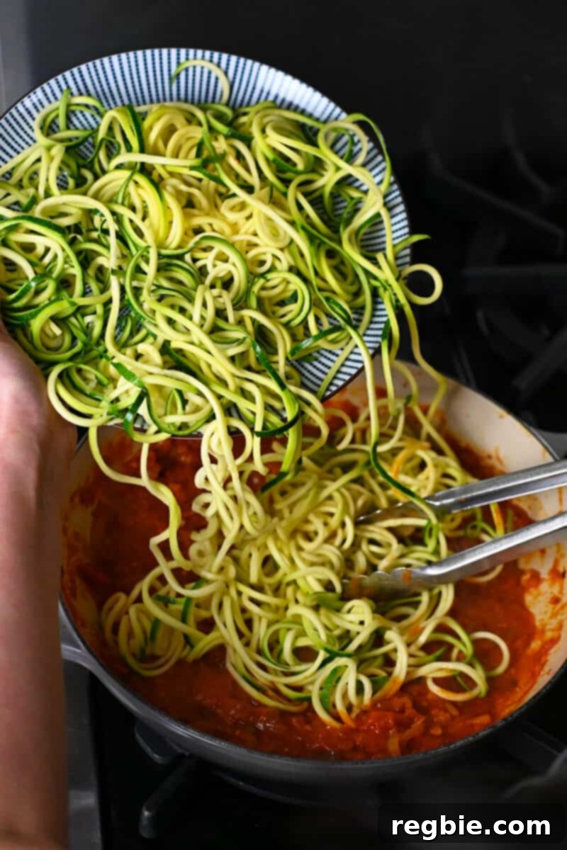 Adding spiralized zucchini to a skillet with hot marinara sauce, preparing for a quick toss.