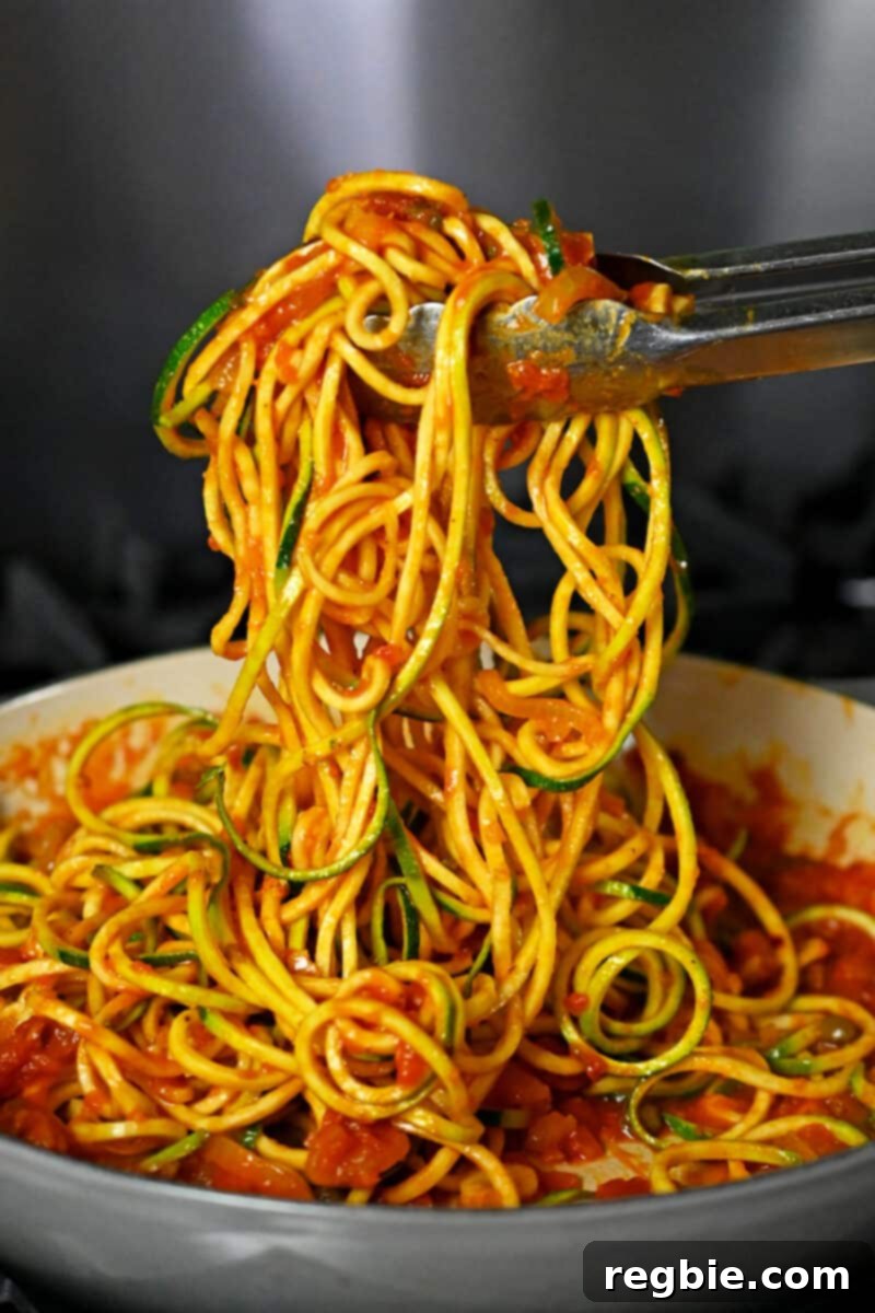 A pair of tongs tossing zucchini noodles in a skillet with spicy marinara sauce, ensuring every strand is coated.