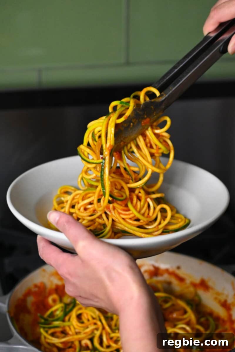 A pair of tongs is transferring zucchini noodles tossed in marinara sauce into a shallow bowl, ready for the shrimp.