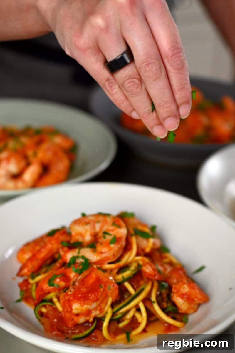 A hand is sprinkling minced Italian parsley on top of a bowl filled with paleo, Whole30, and keto spicy shrimp pasta, adding a final touch of freshness.
