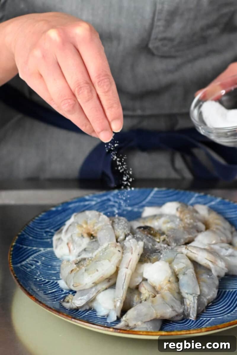 A hand is sprinkling salt on a plate of raw peeled jumbo shrimp, preparing them for cooking.