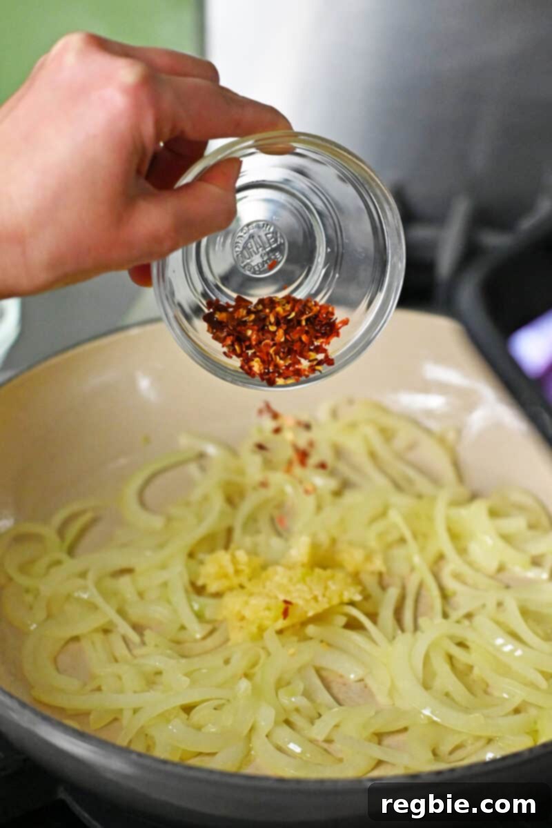 A hand is adding a small amount of red pepper flakes to a pan with sautéed sliced onions and minced garlic, intensifying the aroma.