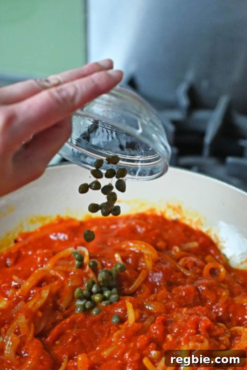 A hand is adding some capers to a skillet filled with marinara sauce, sliced onions, minced garlic, and red pepper flakes, creating a zesty base.
