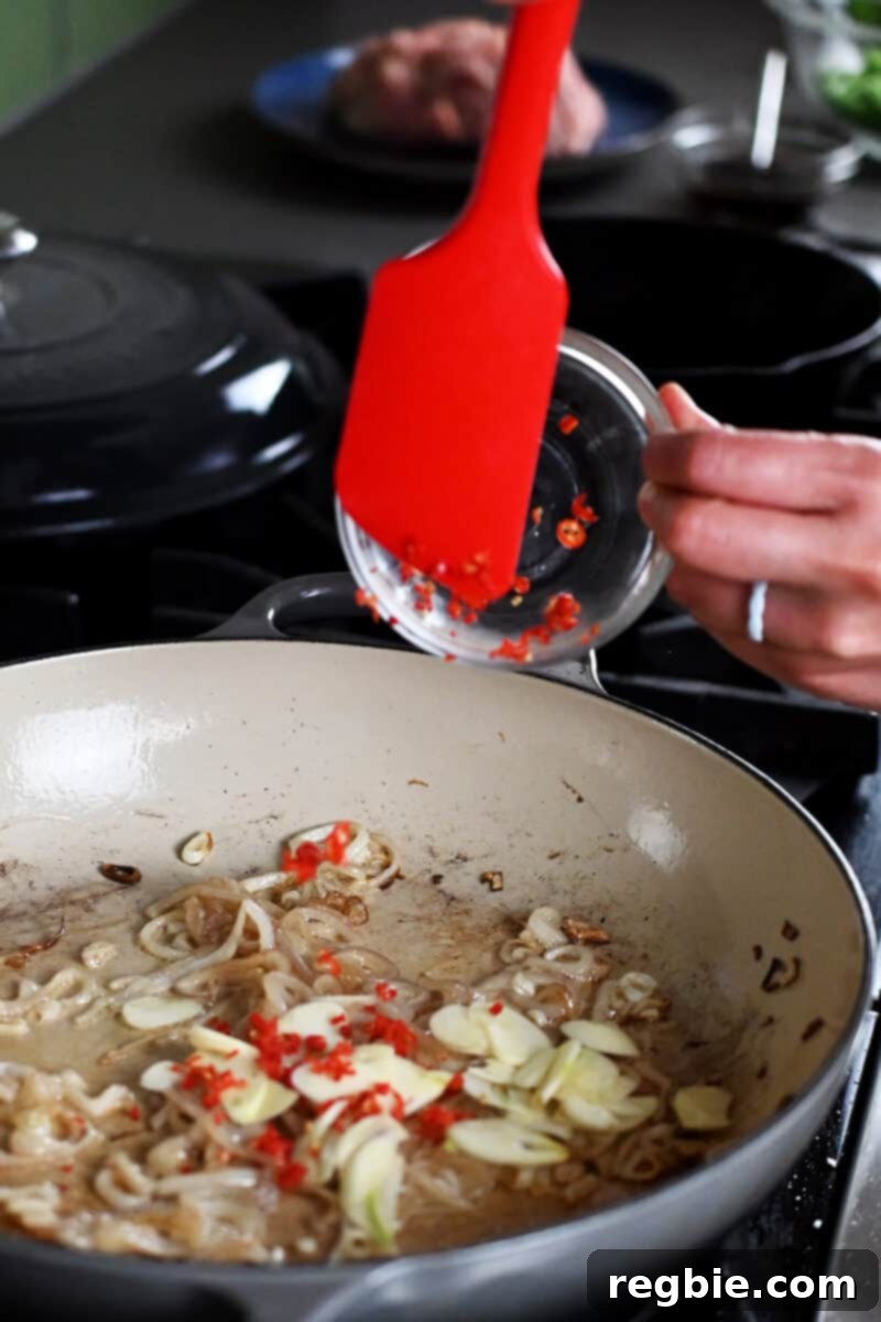 A red spatula is adding sliced garlic and thinly sliced Thai red peppers into a skillet with sautéed shallots.