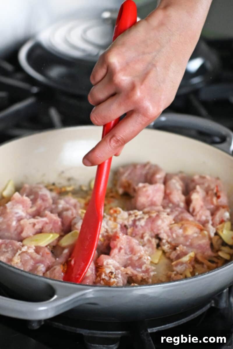 A red spatula is breaking up ground chicken into a skillet to make Thai Basil Chicken