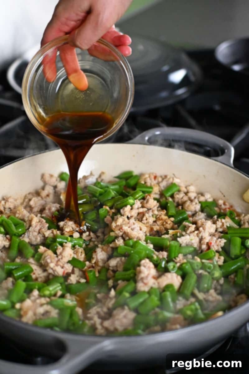 Pouring sauce into a skillet with ground chicken and green beans to make Thai Basil chicken