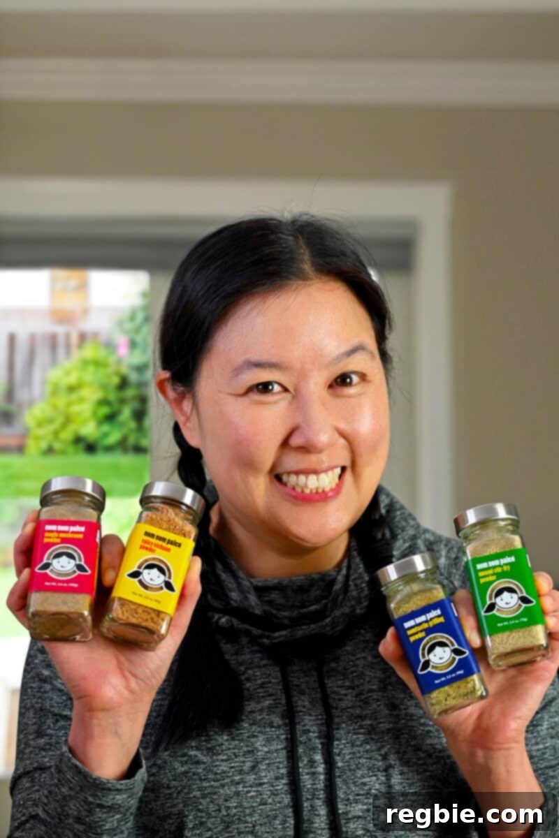 A warm and inviting photograph of Michelle Tam, smiling genuinely as she proudly holds her collection of Nom Nom Paleo spice blend bottles in front of her. The image conveys her personal connection and excitement for the new product line.
