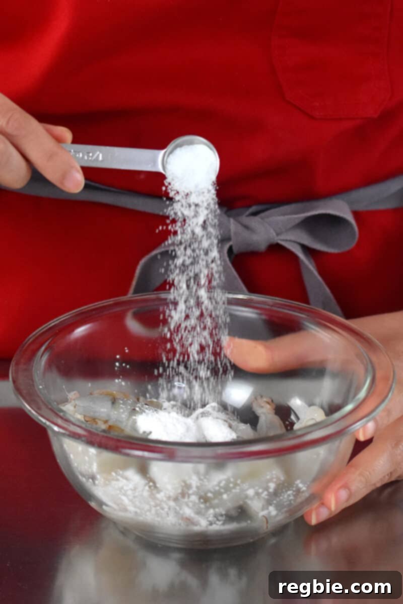 A person in a red apron is adding a spoonful of salt to a bowl of raw shrimp, preparing them for seasoning.