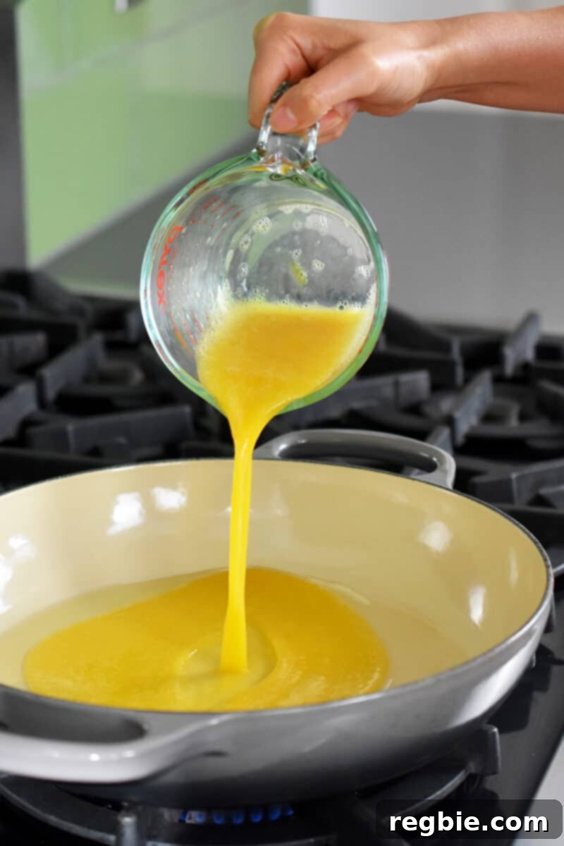 Pouring whisked eggs from a liquid measuring cup into a hot skillet, ready to be scrambled.