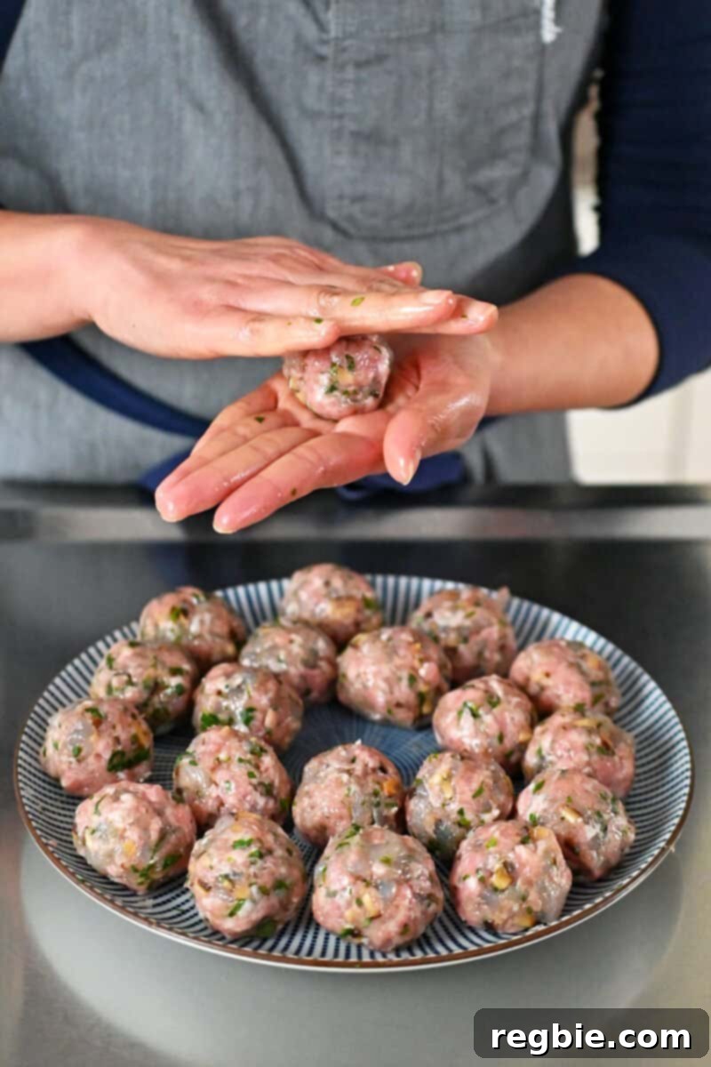 Someone in a gray apron is using their hands, lightly oiled, to roll wonton meatballs and placing them on a blue and white plate.