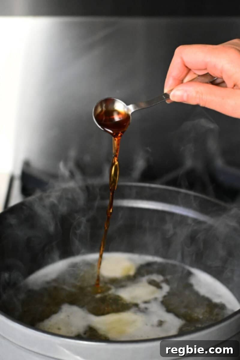 Pouring a spoonful of fish sauce into a simmering pot of chicken broth and ginger, adjusting seasoning.
