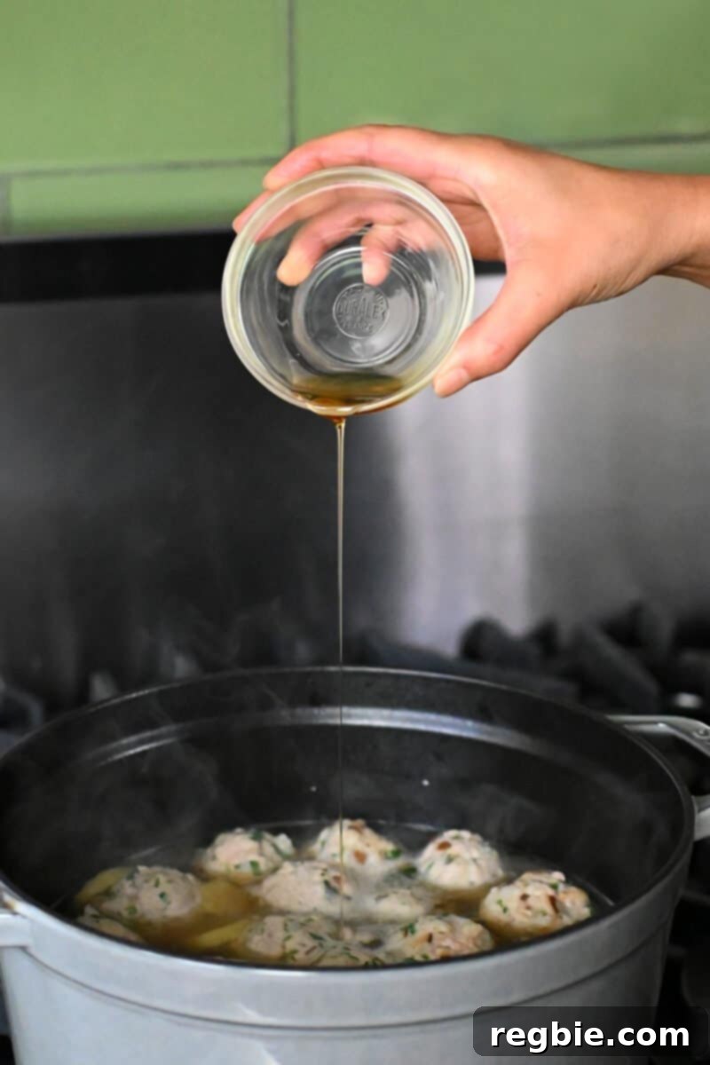 A hand is adding toasted sesame oil from a small bowl to a pot of wonton meatball soup after cooking, for fragrance.