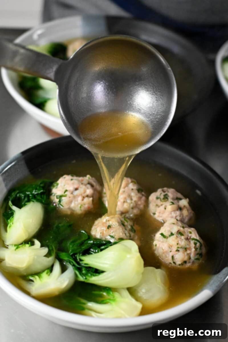 A ladle is adding broth to a bowl filled with wonton meatballs and baby Bok Choy, preparing for garnish.
