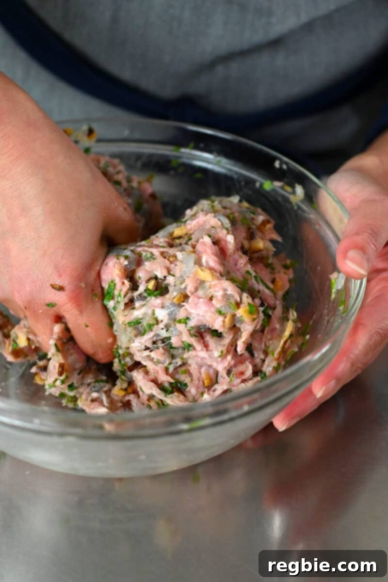 A hand is mixing and squeezing the wonton meatball ingredients in a large glass mixing bowl until well combined.