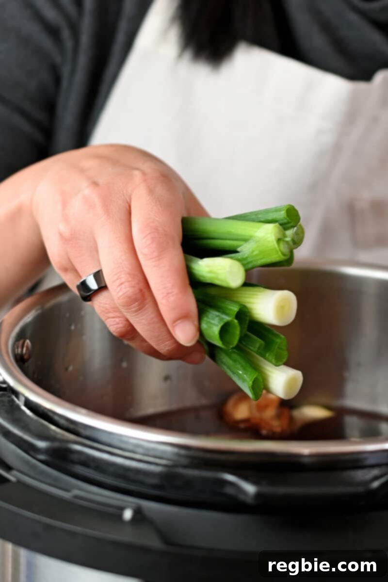 A hand is adding cut scallions to an open Instant Pot, joining the other ingredients for Chinese Beef Noodle Soup.